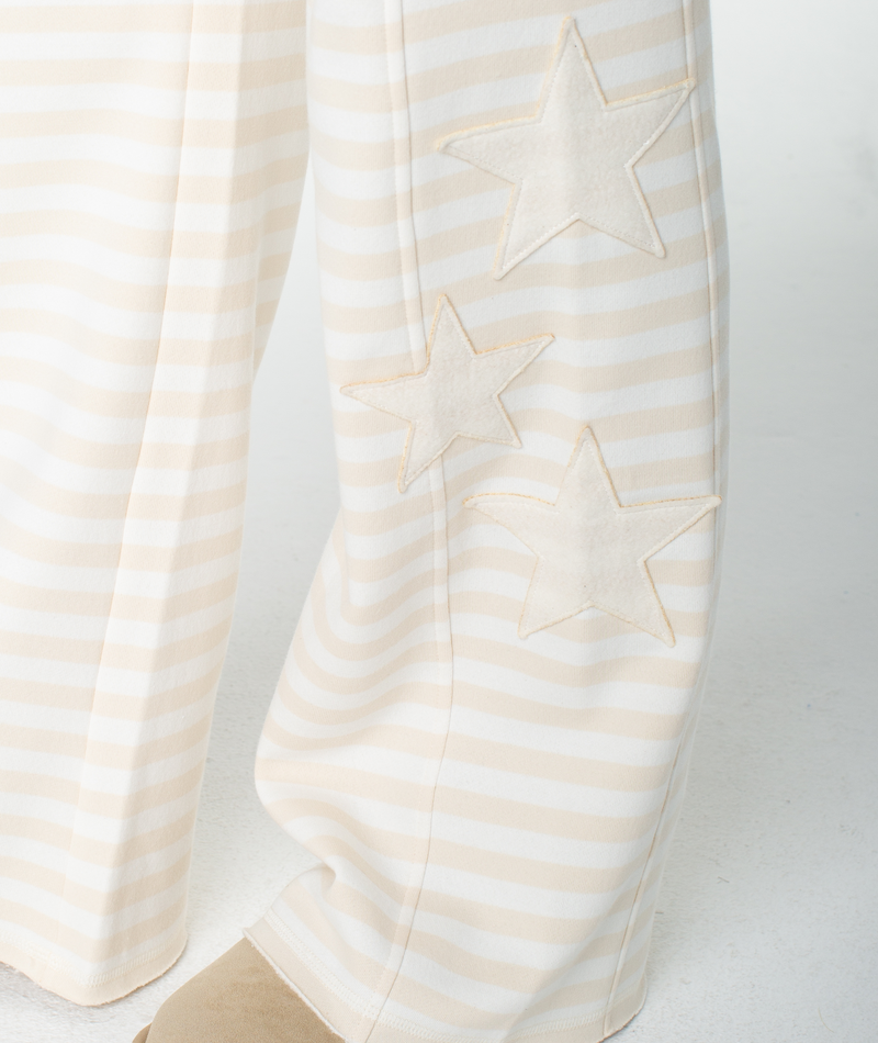 The Fringing Reefs Pant by Roxy features cream and beige stripes with three beige star patches along the side of the leg, making them perfect for relaxing in style on a white surface.