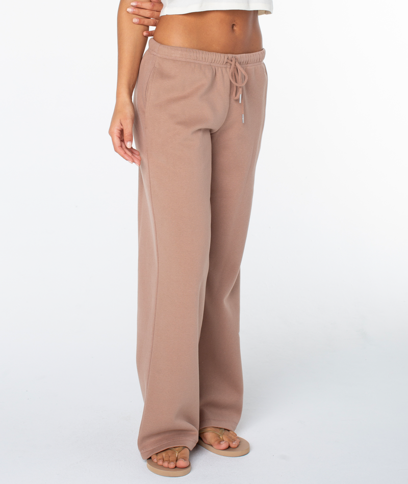 A person wearing loose, light brown Ariel Wide Leg Pant by Roxy with a drawstring waist and open-toed sandals stands against a white background. Only the lower body and part of the upper torso are visible.