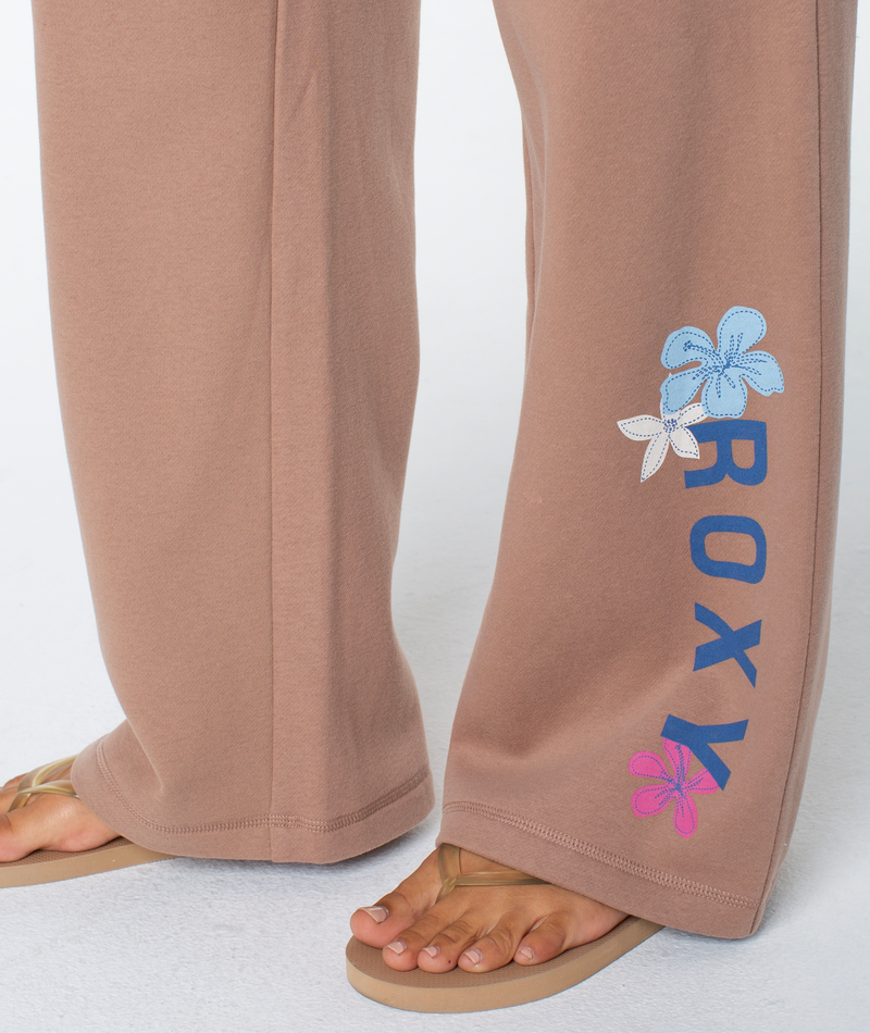 A person wearing the Ariel Wide Leg Pant by Roxy, featuring blue text and floral designs, stands on a white surface in beige flip-flops.