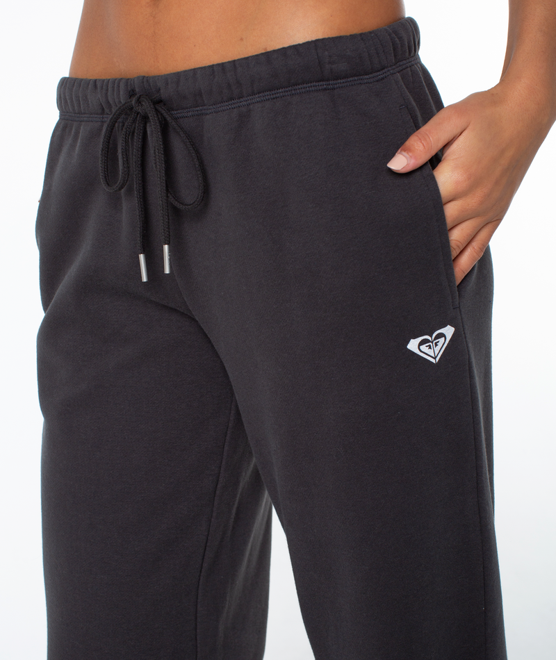 A person wears the Roxy Ariel Wide Leg Pant—black, with a drawstring waist and small white heart logo by the pocket—perfect for beachside lounging, standing against a plain white background.