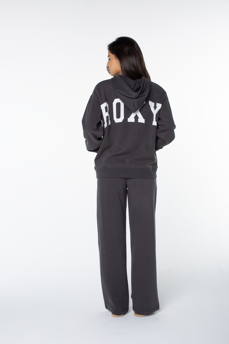 A person stands facing away, wearing a dark cotton blend hooded sweatshirt with ROXY in large white letters on the back and matching Ariel Wide Leg Pant, against a plain white background.