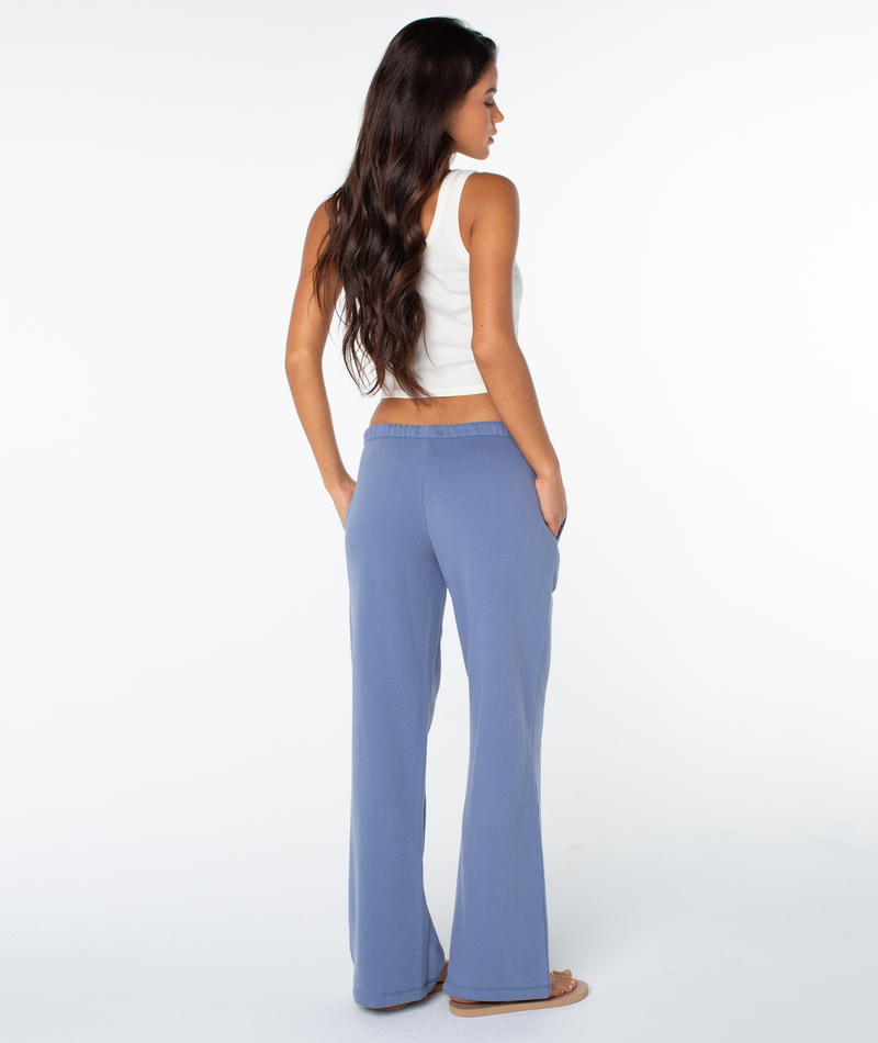 A woman with long brown hair stands facing away, hands in the pockets of blue Ariel Wide Leg Pant by Roxy, styled with a white sleeveless top and sandals against a plain white background.