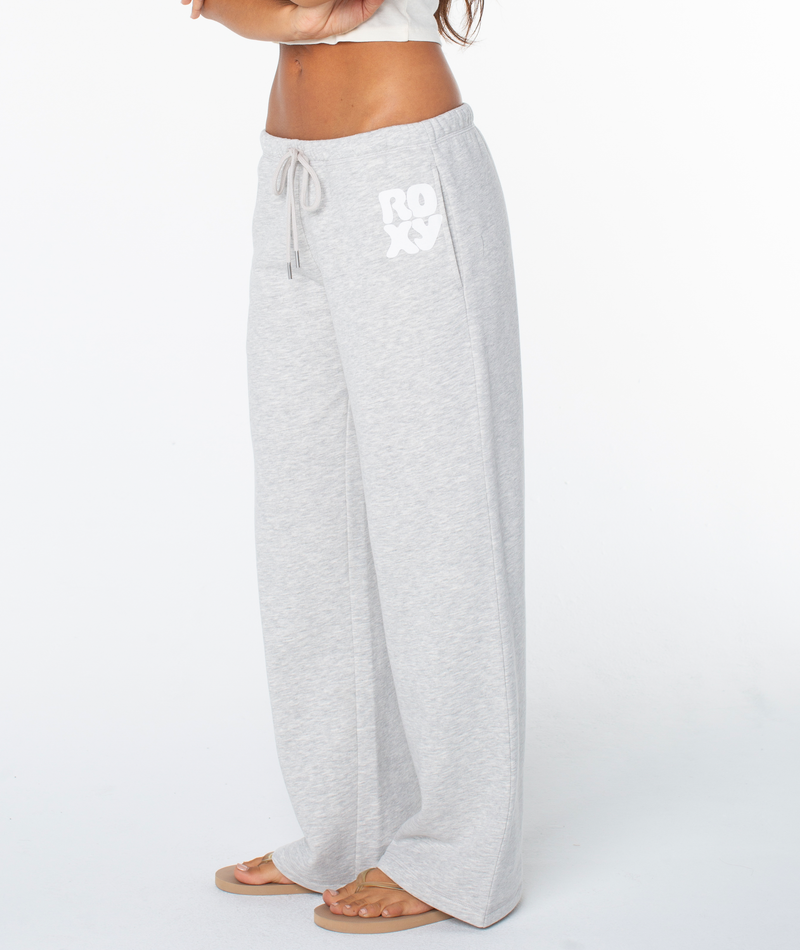 A person in light gray Ariel Wide Leg Pant by Roxy, featuring a soft cotton blend and ROXY on the pocket, pairs them with a white crop top and sandals against a white background.