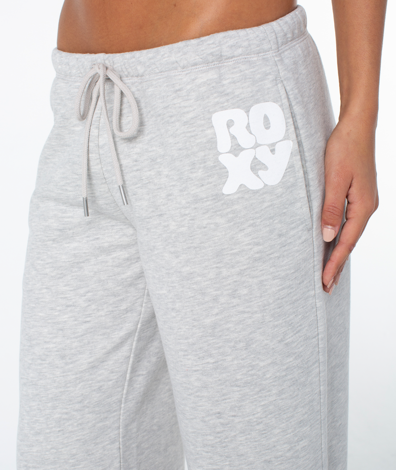 Person wearing light gray Ariel Wide Leg Pant by Roxy with ROXY in white on the left side, featuring a cotton blend, drawstring waist, and comfy wide leg fit. One hand rests on their thigh.