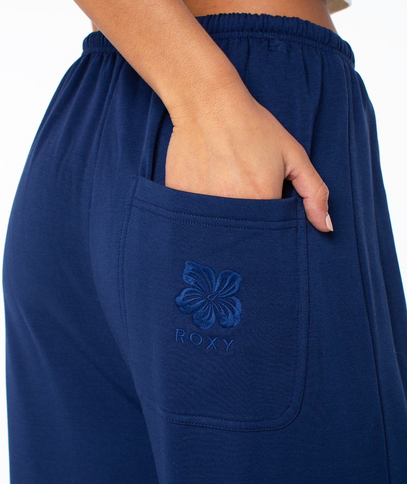 Close-up of a hand resting in the back pocket of navy blue Roxy Surfing By Moonlight Pant, featuring an embroidered flower and logo for all-day comfort.