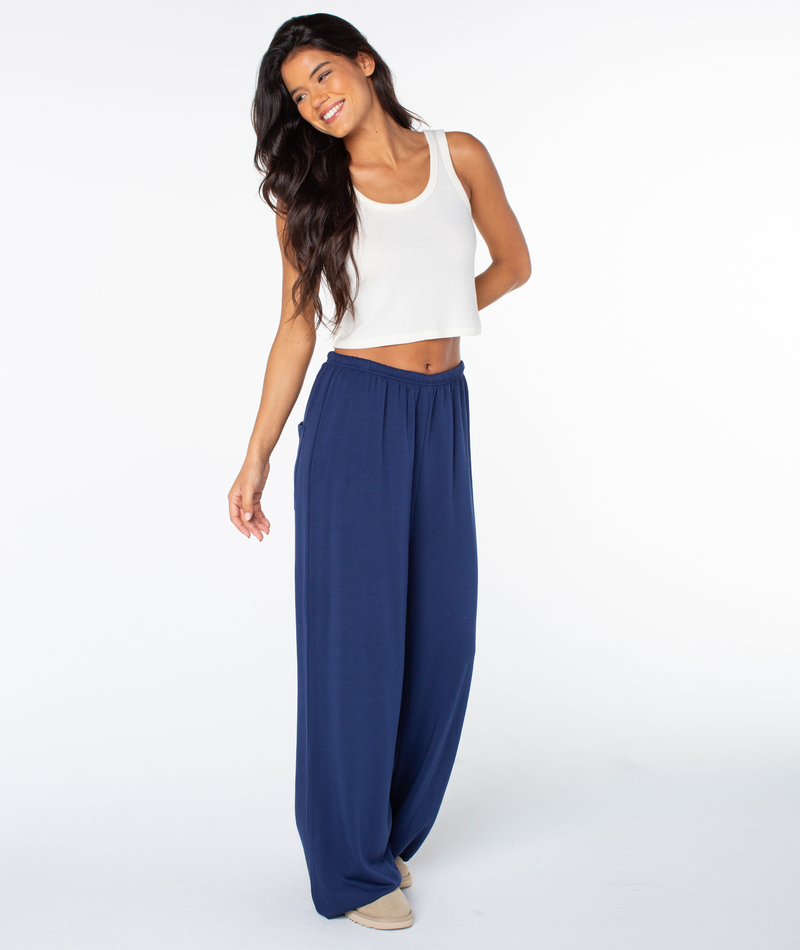 A person wearing a white tank top and flattering, wide-leg Surfing By Moonlight pants in Naval Academy Hibiscus Emb.