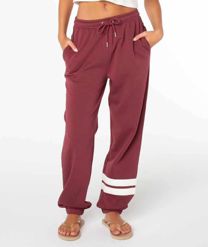 A person is seen standing barefoot in flip-flops against a plain white background, wearing Roxy’s Sunset Pant joggers in maroon, which feature two distinctive white stripes near the left ankle. The joggers are made from a comfortable cotton blend for all-day ease, with hands casually tucked into their pockets.