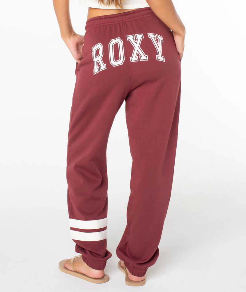 A person wearing the Roxy Sunset Pant in maroon—a soft cotton blend jogger sweatpant featuring “ROXY” in white letters on the back and two white stripes near the left ankle—stands barefoot on a white background.
