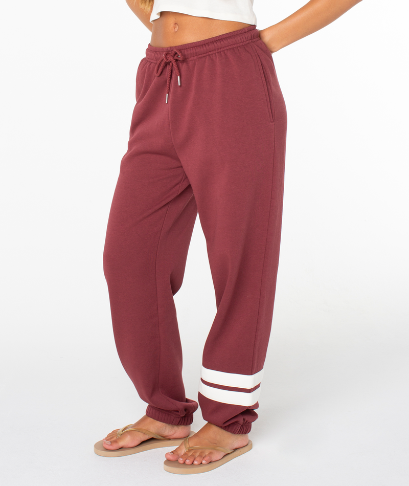 A person wearing the Roxy Sunset Pant in a maroon cotton blend with white stripes near the ankle, paired with a white top and beige flip-flops, stands against a plain white background.