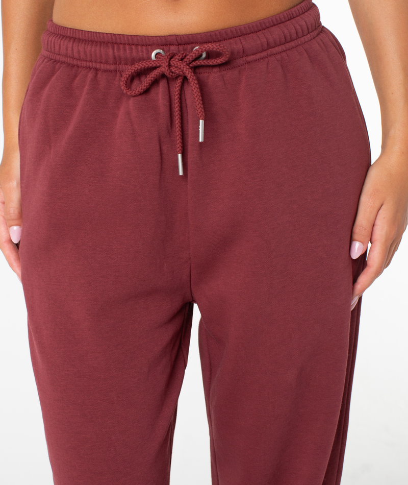 A person wearing the Roxy Sunset Pant in maroon, featuring a drawstring waistband and made from a soft cotton blend, stands against a plain white background with only their lower torso and upper legs visible.