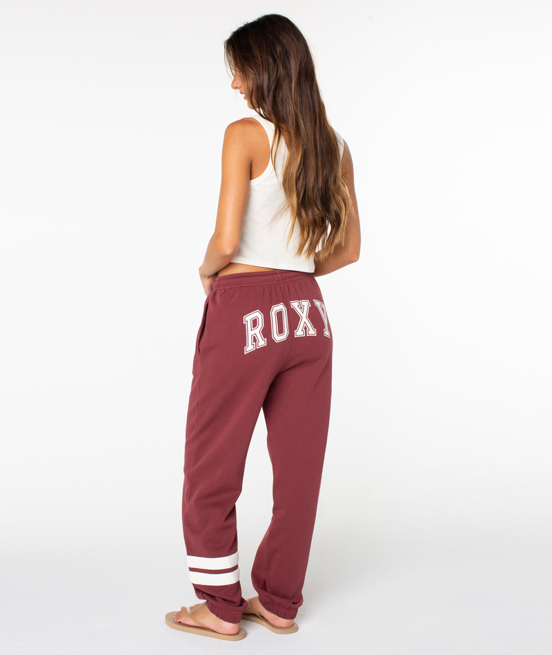 A woman stands facing away, wearing a white tank top and the Roxy Sunset Pant, maroon cotton-blend jogger sweatpants with “ROXY” on the back and white stripes near the cuffs, paired with flip-flops. She is set against a plain white background.