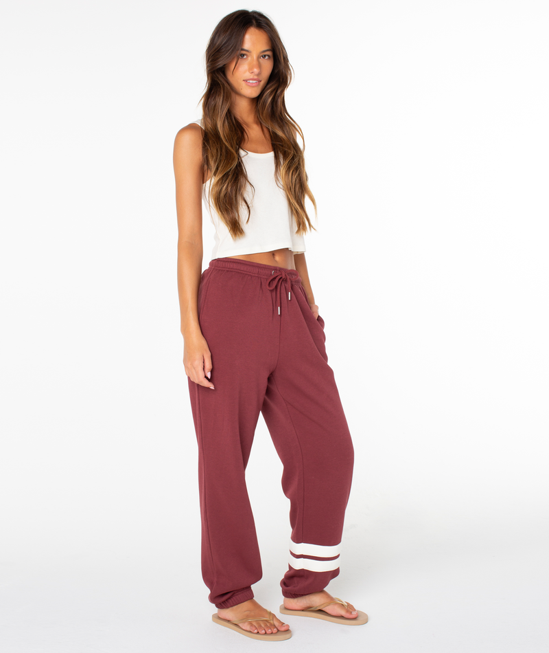 A woman with long brown hair wears a white crop top and the Roxy Sunset Pant in Port End Game—a maroon jogger sweatpant with white stripes near the ankle crafted from a soft cotton blend—paired with beige flip-flops. She stands on a white background, looking at the camera.
