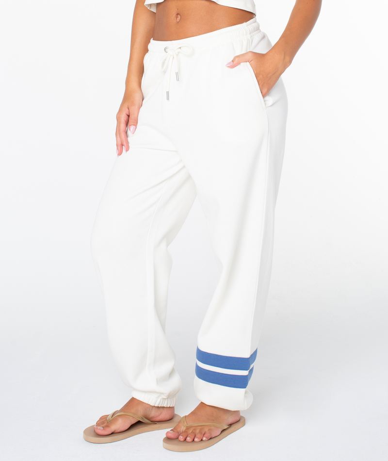 A person stands in flip-flops, wearing Roxy's Sunset Pant—white cotton blend jogger sweatpants with two blue ankle stripes, side pockets, and an elastic waistband. The upper body is mostly out of frame.