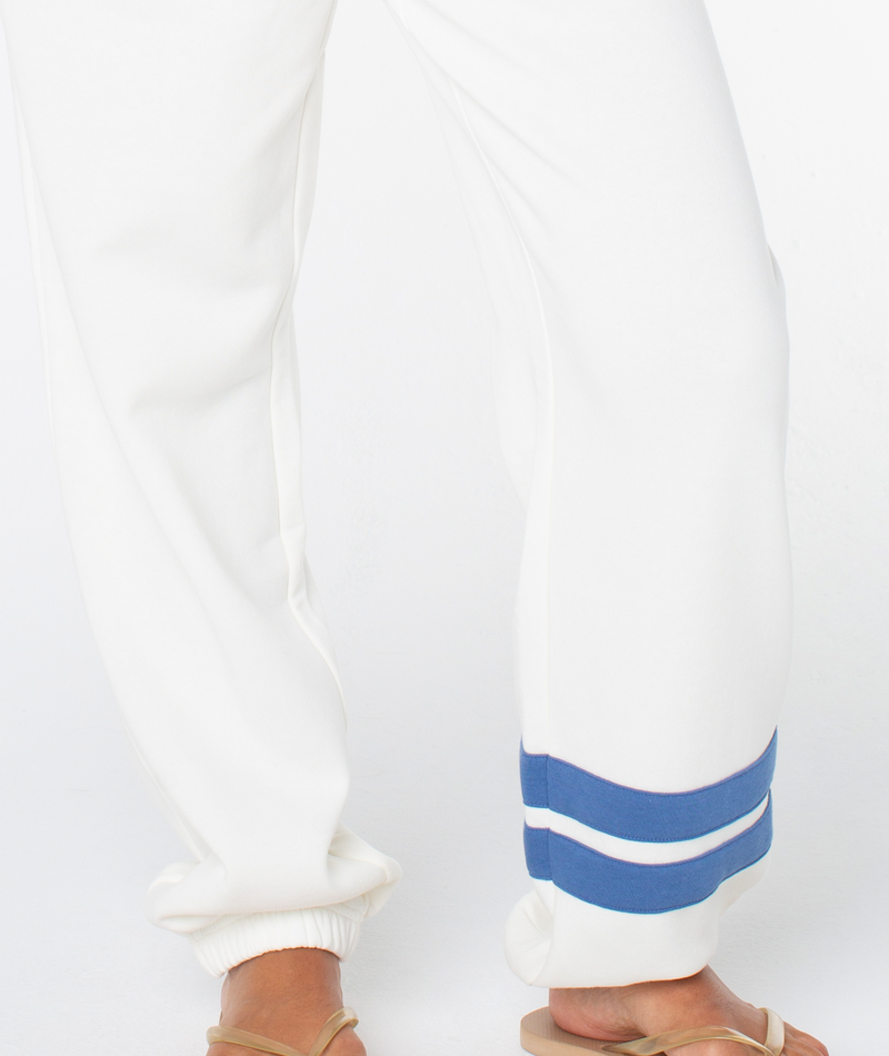A person wearing loose white Roxy Sunset Pant with two blue stripes near the bottom hem is standing barefoot on a white background.