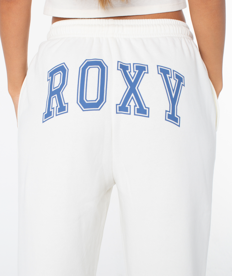 A person models the Roxy Sunset Pant—white cotton blend joggers with "ROXY" in bold blue lettering on the back, hands in pockets, against a plain white background.