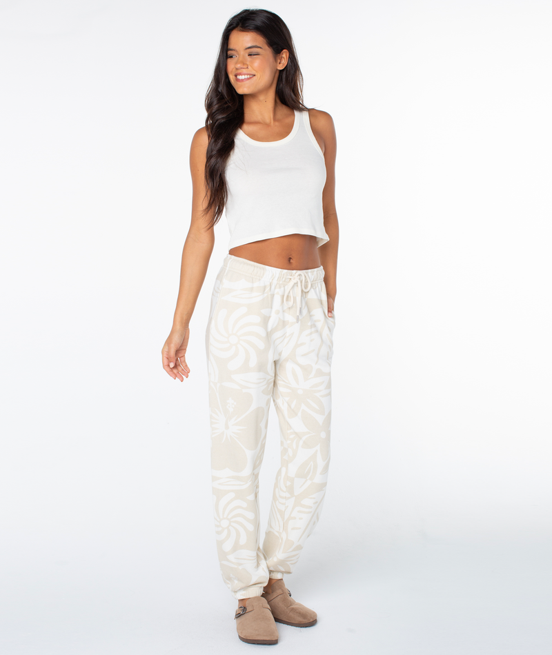 A person wears a white cropped tank top and Roxy Maile Printed Pant in parchment cool surf featuring a large-scale white floral print, paired with beige slide sandals.