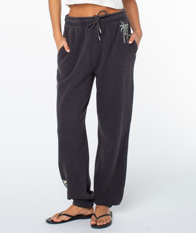 A person wears loose black Roxy Maile Pant sweatpants with pockets, palm tree embroidery on the hip, and black flip-flops. Only their lower half is visible against a plain white background.
