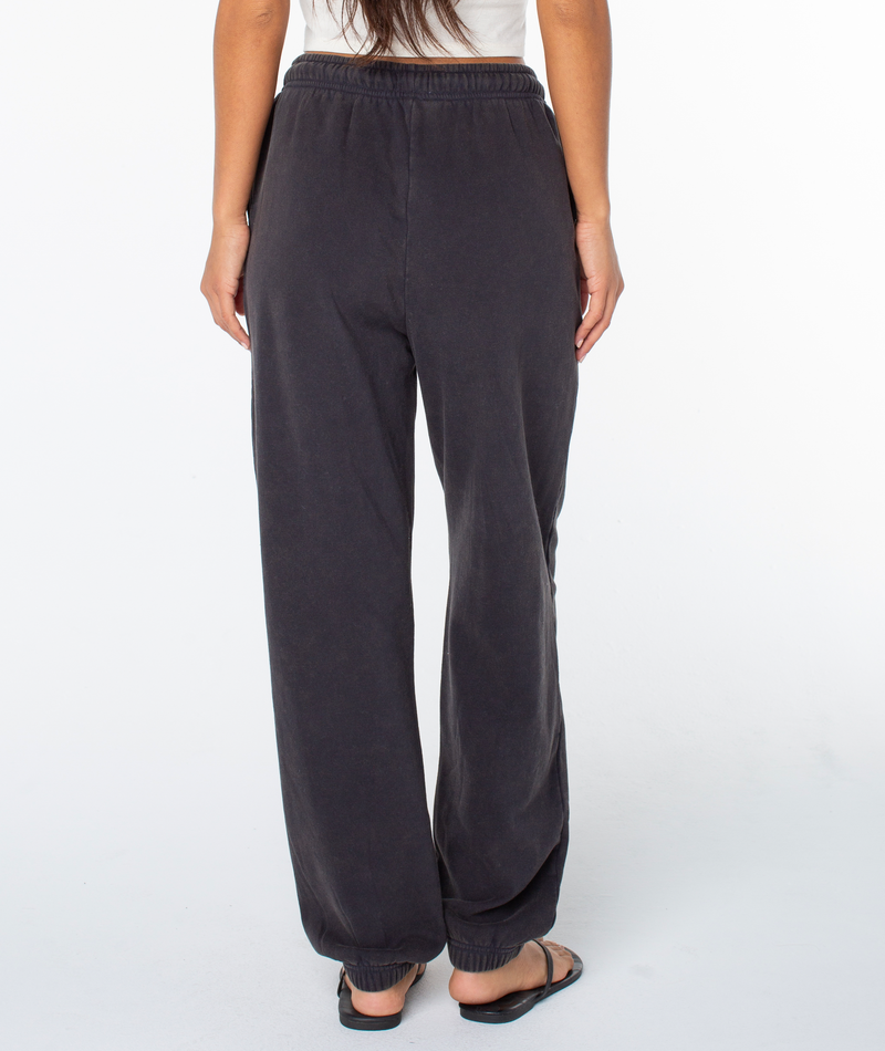 A person faces away, wearing a white top and Roxy Maile Pant in cozy cotton blend black with an elastic waistband and black slide sandals, set against a plain white background.