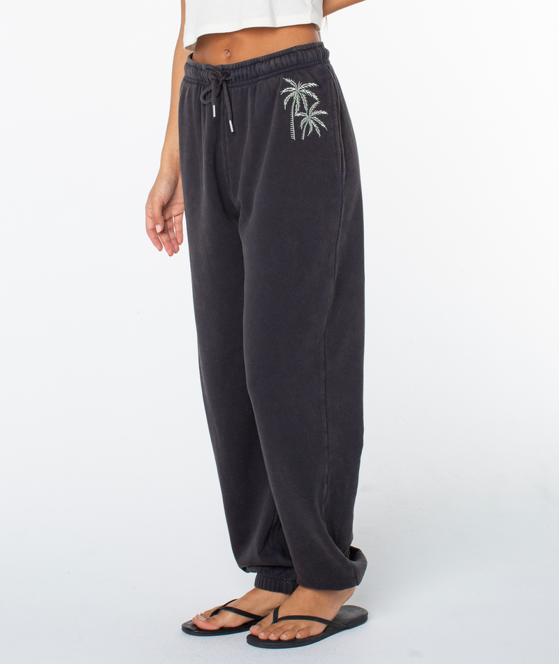 Woman wearing Roxy Maile Pant—black sweatpants in a cozy cotton blend with embroidered palm trees—with a white crop top and black flip-flops, standing against a plain white background (shoulders down visible).