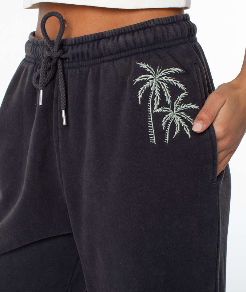 A close-up of a person wearing the Roxy Maile Pant, featuring an embroidered palm tree design near the right pocket, cozy cotton-blend fabric, drawstring waist, and a hand tucked in the pocket.