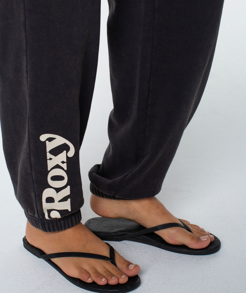 Someone wearing black Maile Pant by Roxy, made from a cozy cotton blend, and black flip-flops stands on a light surface.