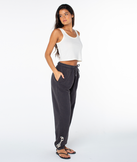 Woman standing in a relaxed pose, wearing a white crop tank top, Roxy Maile Pant in Phantom Palm Tree Resort with chain detail near the ankle, and flip-flops, against a plain white background.