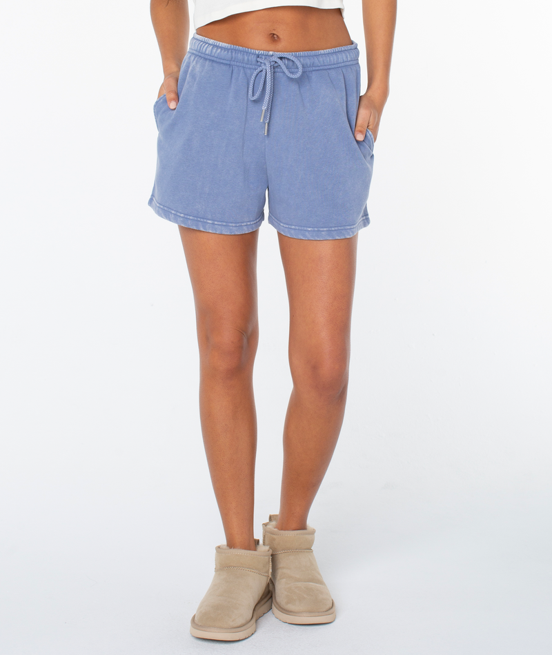 A person wears blue Roxy Hazy Daze Short in a soft cotton blend with tan slippers, hands in pockets, against a plain white background; only their lower torso and legs are visible.