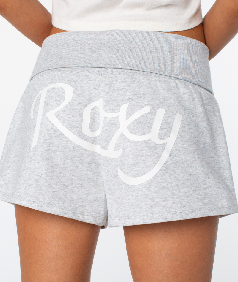 A person wearing the Roxy Sunday Short in light gray, featuring “Roxy” in white script across the back, shown from waist to mid-thigh, with part of a white top visible.