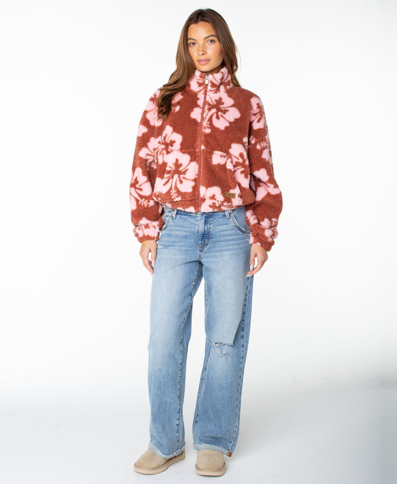 A woman stands against a white background wearing the Roxy Cold Surf Zip Up in Chutney Hibiscus, styled with light blue wide-leg jeans and beige slip-on shoes.