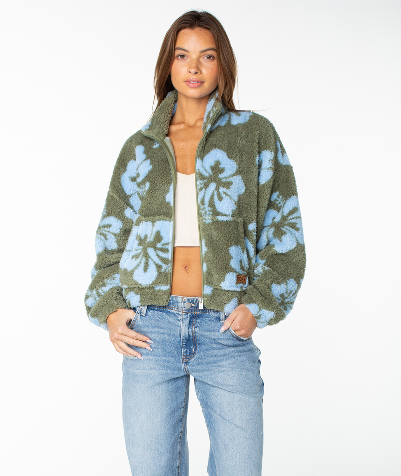 A woman stands against a plain white background wearing the Roxy Cold Surf Zip Up—a green fleece jacket with a hibiscus print—layered over a white cropped top and paired with light blue jeans.