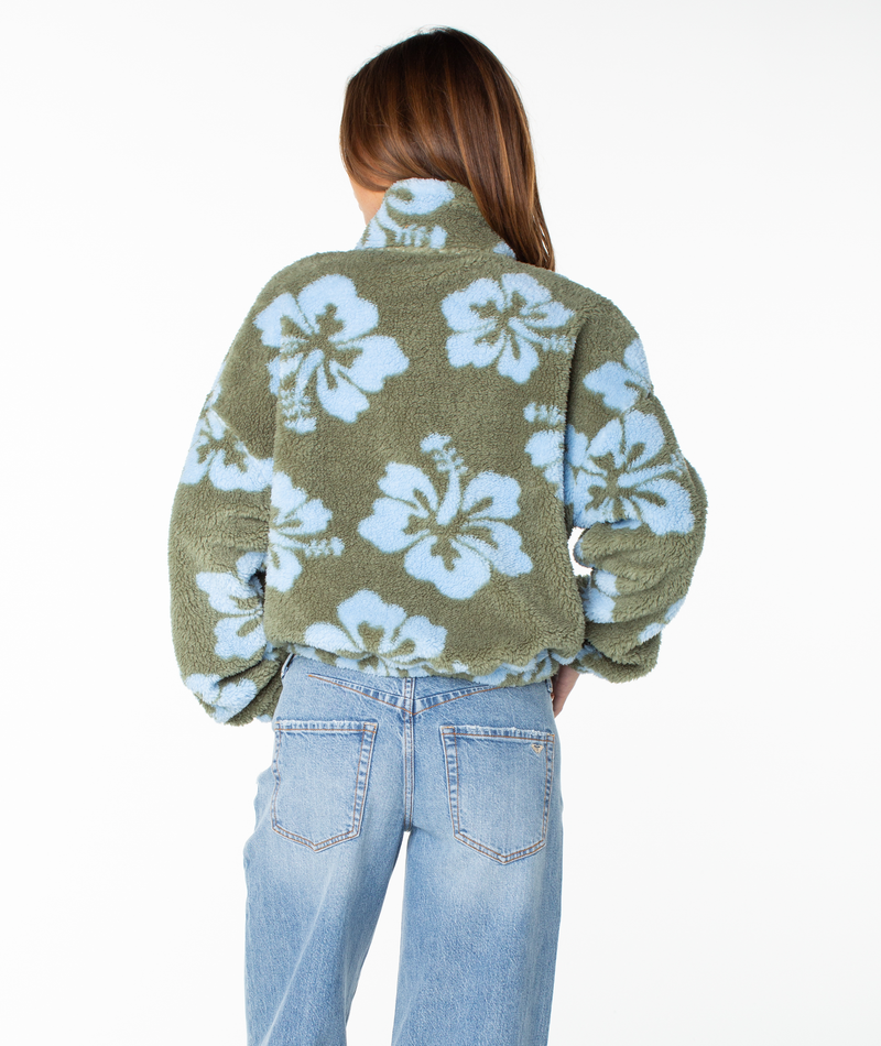 A person stands facing away, wearing light blue jeans and the Roxy Cold Surf Zip Up—a green fleece jacket with a cropped fit and a bold, oversized light blue hibiscus print—against a plain white background.