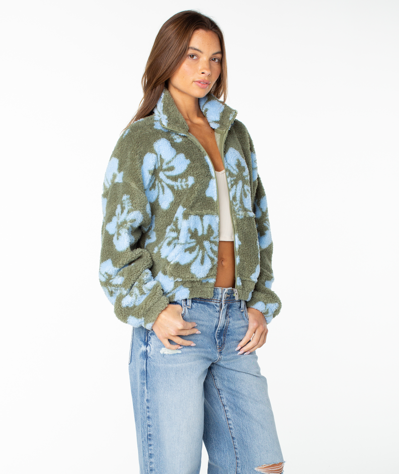 A person wearing the Roxy Cold Surf Zip Up in Oil Green Hibiscus, styled with a white crop top and light ripped jeans, stands against a plain white background.