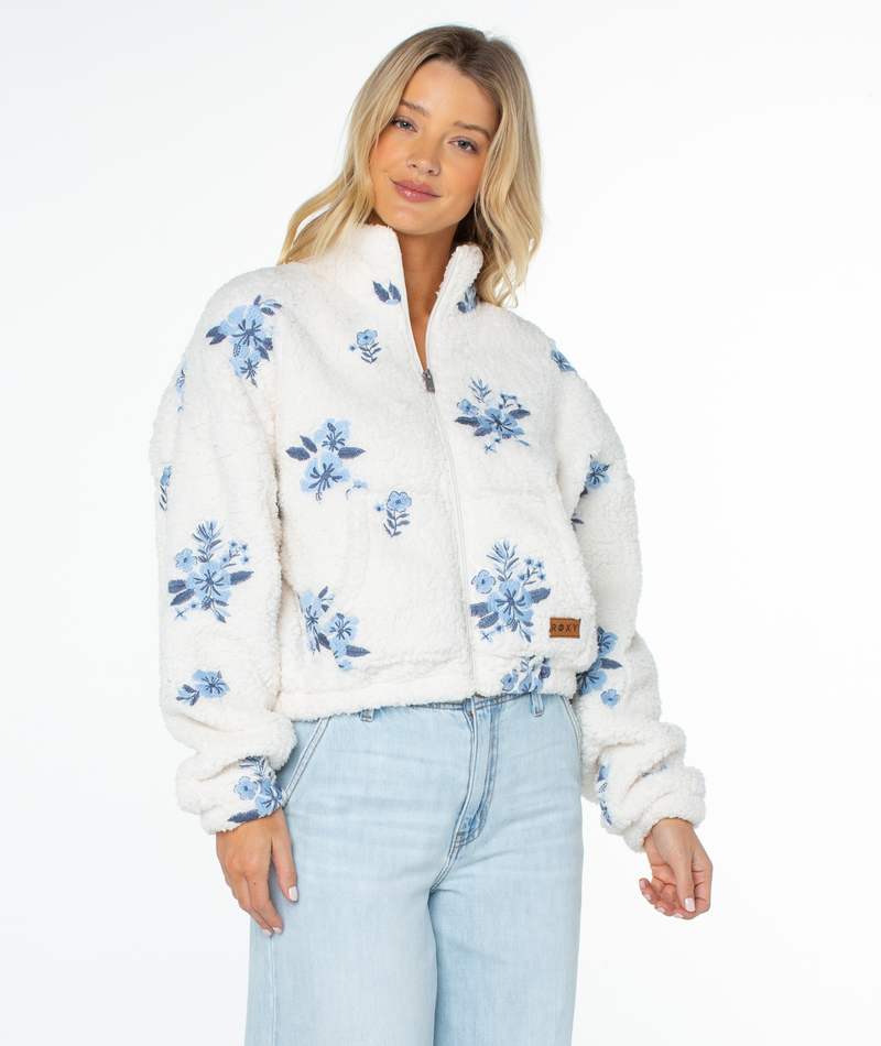 A woman wears a white, floral-patterned Roxy Cold Surf Fleece Top featuring a relaxed silhouette, high collar, and front zipper.