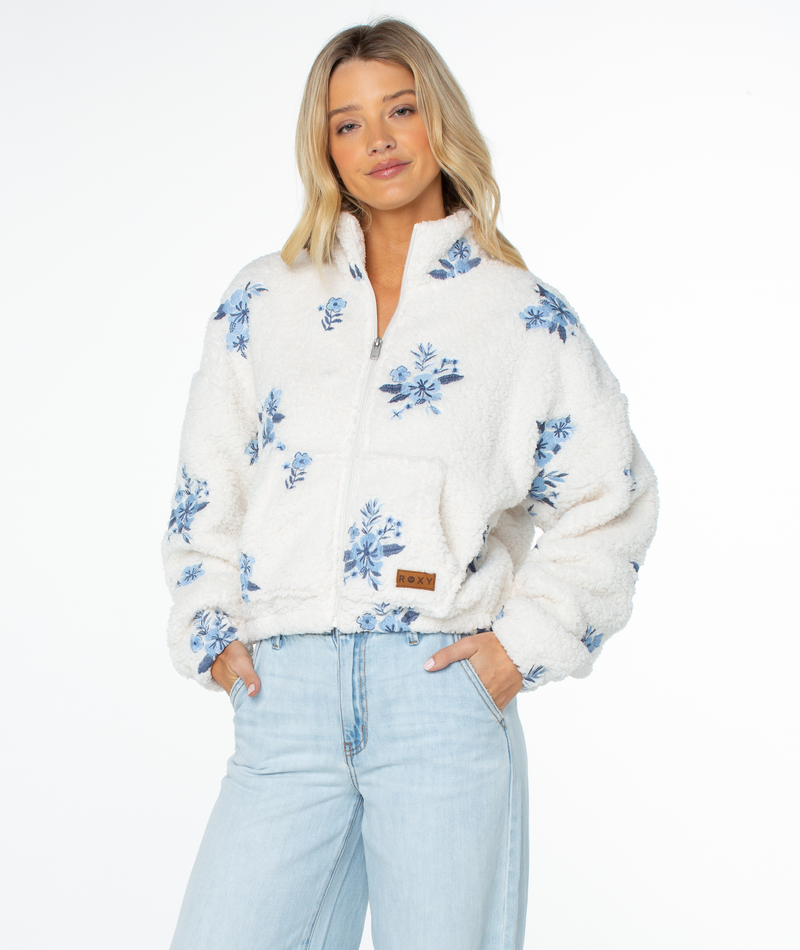 A woman wears a white, fuzzy, zip-up Roxy Cold Surf Fleece Top featuring a blue floral print, paired with light-wash jeans for a relaxed silhouette in cooler weather.
