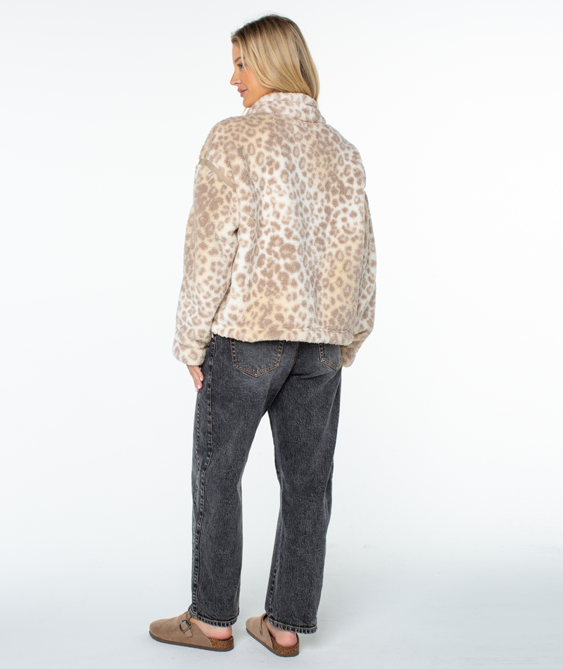 A woman stands facing away, wearing a beige leopard-print Roxy Wurlie Fleece Jacket, dark grey jeans, and tan slip-on shoes.
