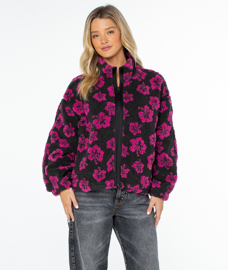 A person wears a black, zip-up Roxy Wurlie Fleece Jacket featuring a bright pink floral print, paired with grey jeans for a coastal cool look.