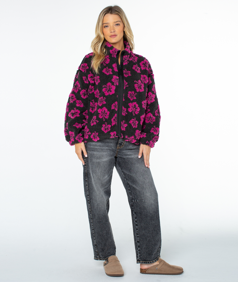A person wearing a Roxy Wurlie Fleece Jacket in Phantom Jacquard Hibiscuss, styled with dark gray jeans and tan slip-on shoes for a coastal cool look.