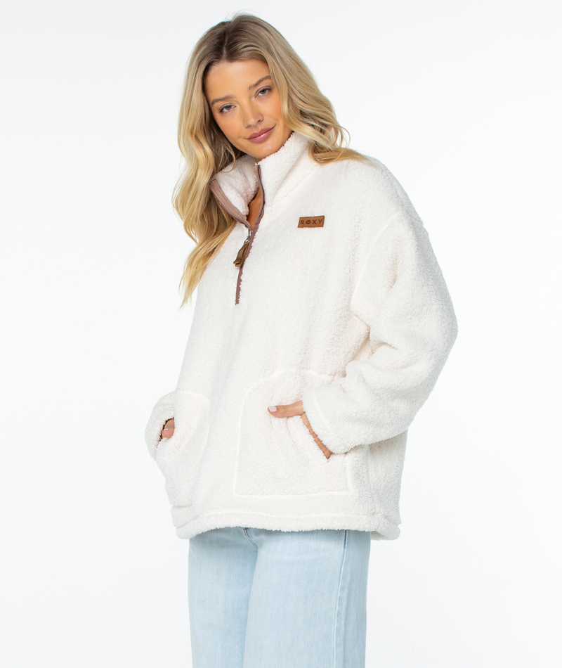 A person wears an off-white Roxy Cozy Day Fleece Top, an off-white, long-sleeve fleece pullover with a cozy mock neck, partial zipper, and oversized kangaroo pocket.
