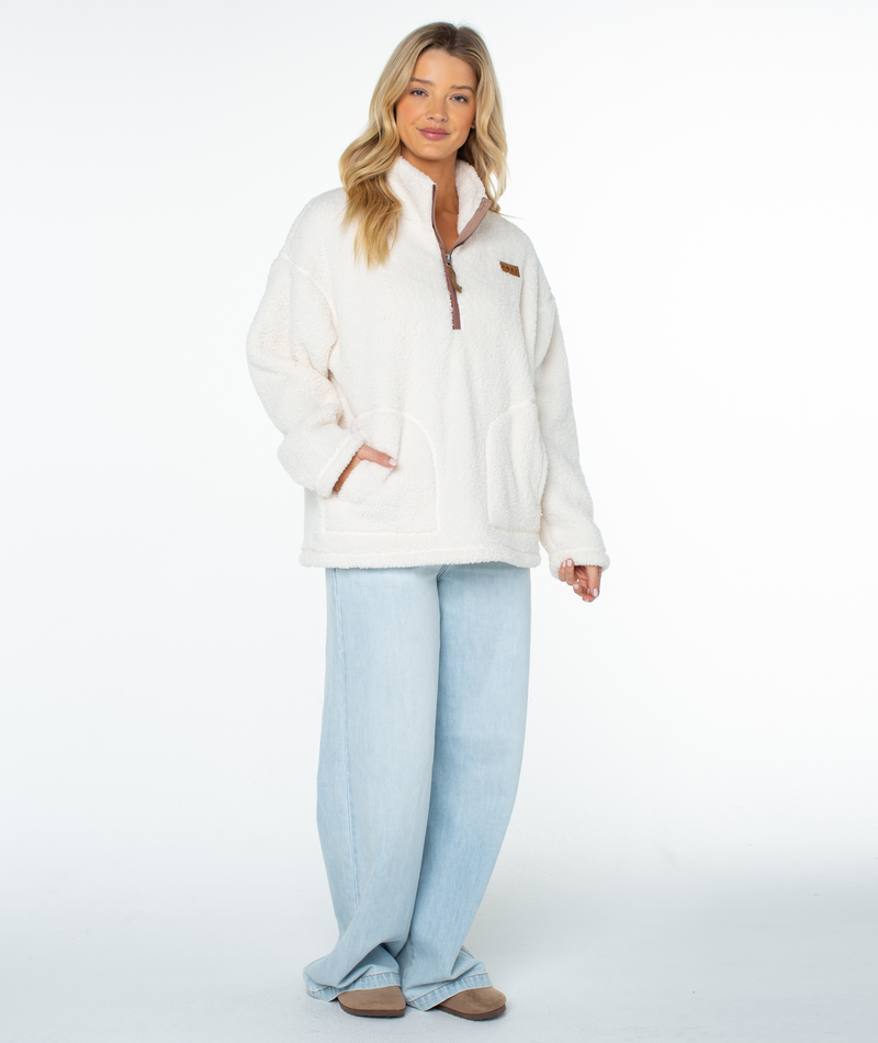 A person wearing a Roxy Cozy Day Fleece Top in off-white, featuring a cozy mock neck and a kangaroo pocket, paired with light-wash wide-leg jeans.