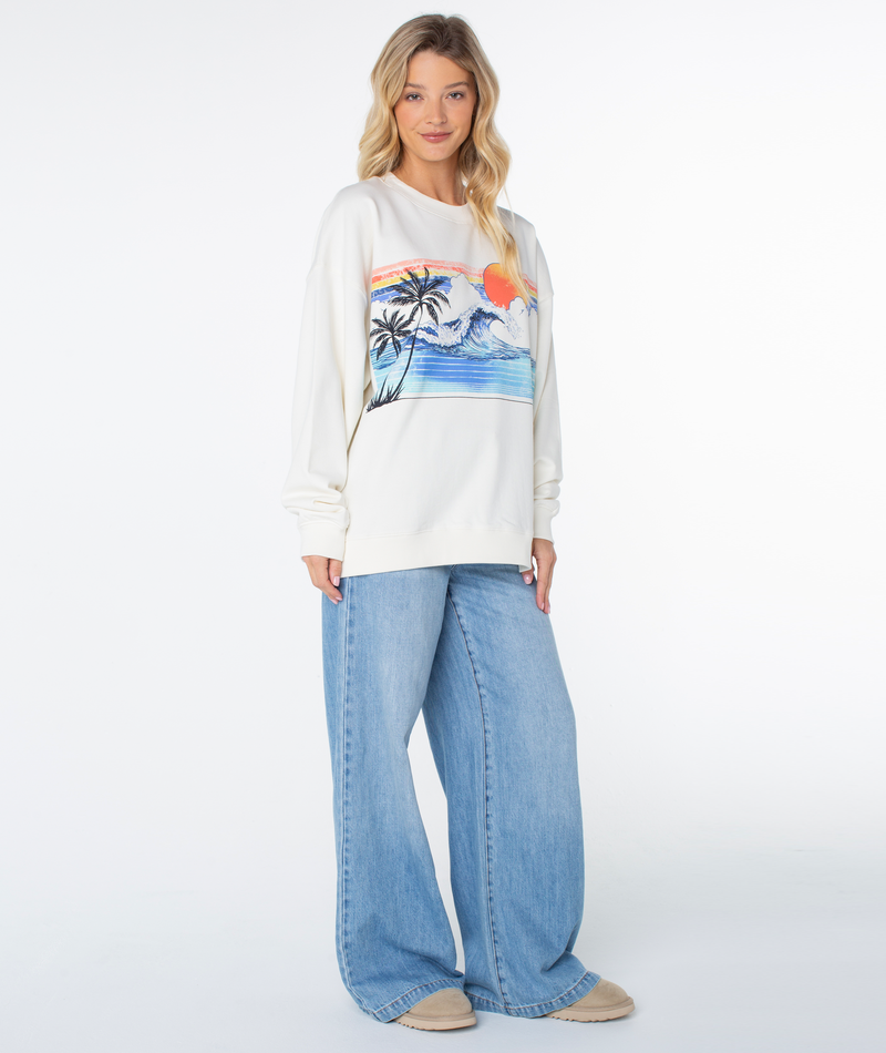 A person wears a Coconut Milk Wave Roxy Line Up Crew Top featuring a graphic print of an ocean wave, palm trees, and a sunset, paired with wide-leg blue jeans for coastal comfort.