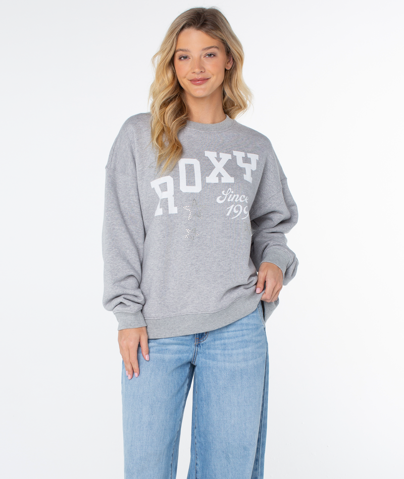 Woman with long blonde hair wearing a grey Line Up Crew Top by Roxy and light blue jeans stands against a plain white background, smiling with one hand in her pocket.