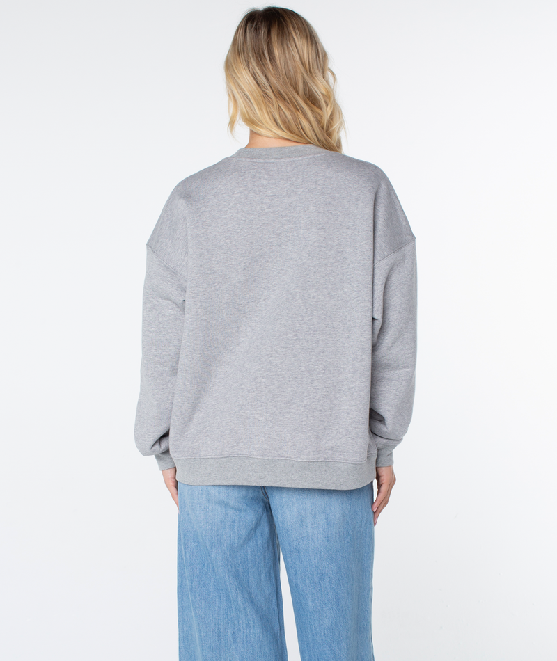 A person with long blonde hair wears the Roxy Line Up Crew Top in gray and light blue jeans, standing with their back to the camera against a plain white background.
