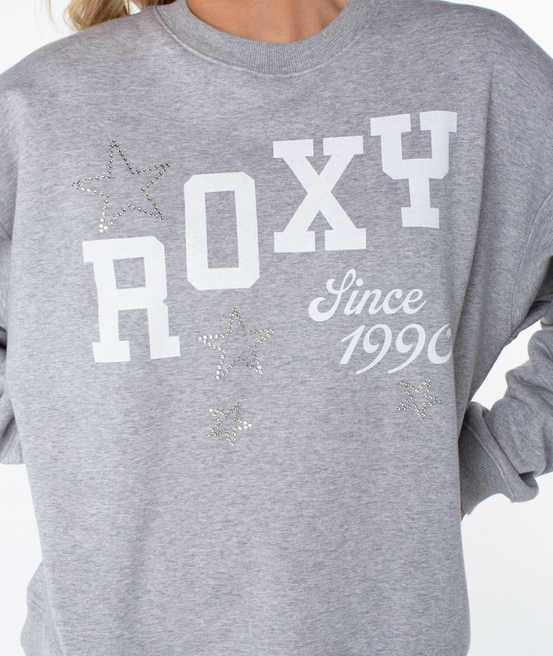 Wearing the gray Roxy Line Up Crew Top with ROXY Since 1990 and rhinestone star designs, capturing effortless coastal comfort; only the torso is visible, head out of frame.