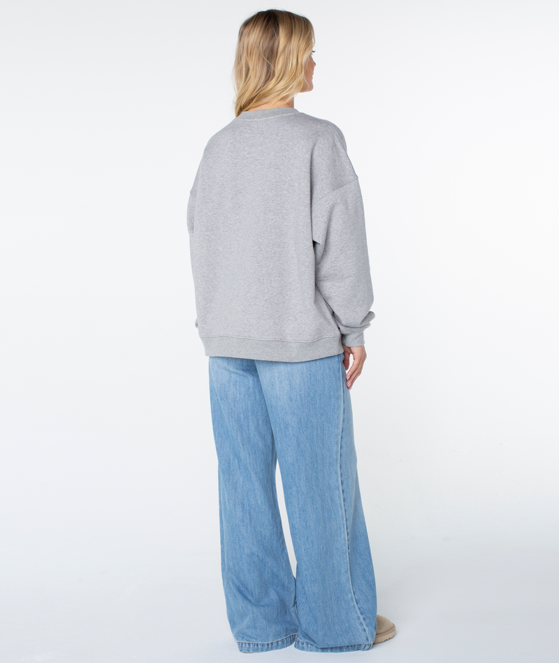 A person with long blonde hair stands facing away, wearing a loose gray Line Up Crew Top by Roxy, wide-leg blue jeans, and beige shoes, radiating coastal comfort against a plain white background.