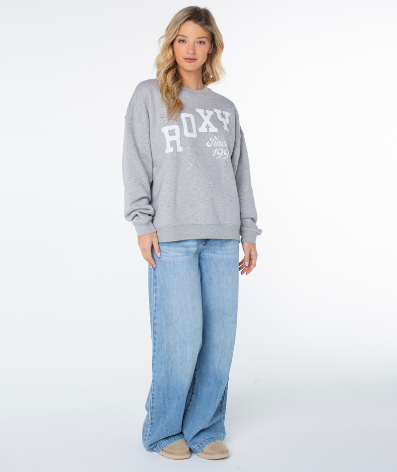 A woman wears the Roxy Line Up Crew Top in Heather Grey High Tides, paired with wide-leg light blue jeans and beige shoes, standing against a white background and looking at the camera.