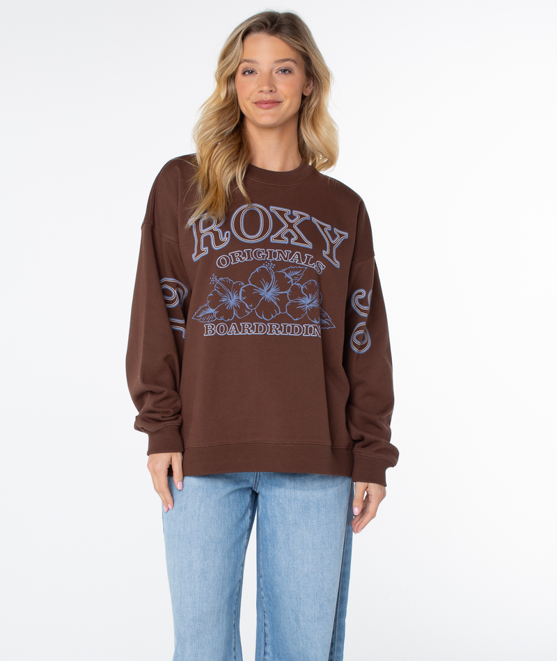 A woman wears a brown Roxy Line Up Crew Top featuring a blue graphic of hibiscus flowers and text on the front and sleeves for coastal comfort.