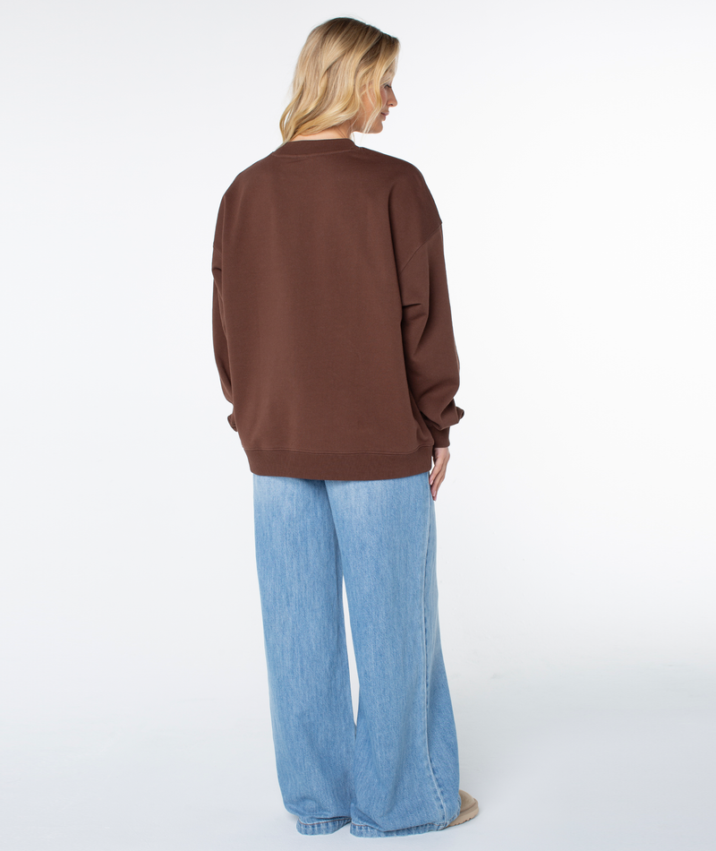A person seen from behind wears a brown Roxy Line Up Crew Top and light-wash blue jeans for coastal comfort.