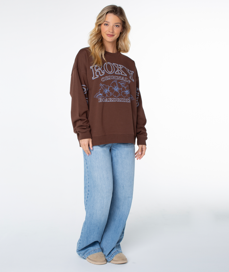 A person wearing a chestnut Roxy Originals Line Up Crew Top with blue floral and text graphics, paired with light-wash wide-leg jeans for coastal comfort.