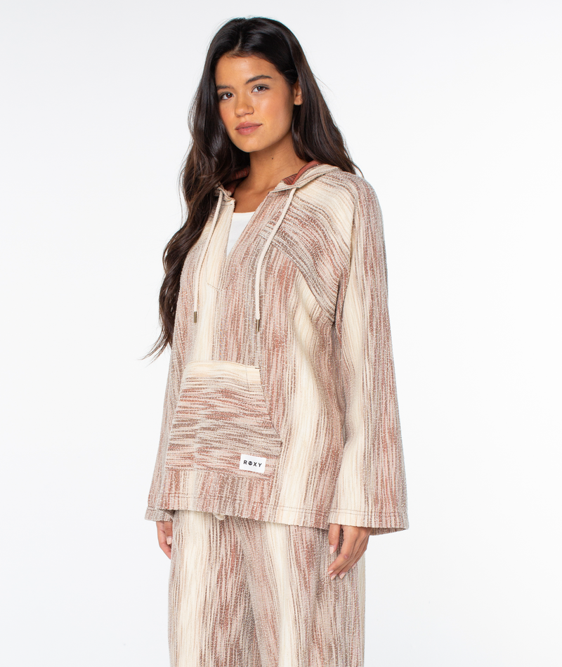 A woman wears the Roxy Wild And Free Top, a relaxed-fit, hooded pullover with a tan and white textured striped pattern and a front kangaroo pocket.