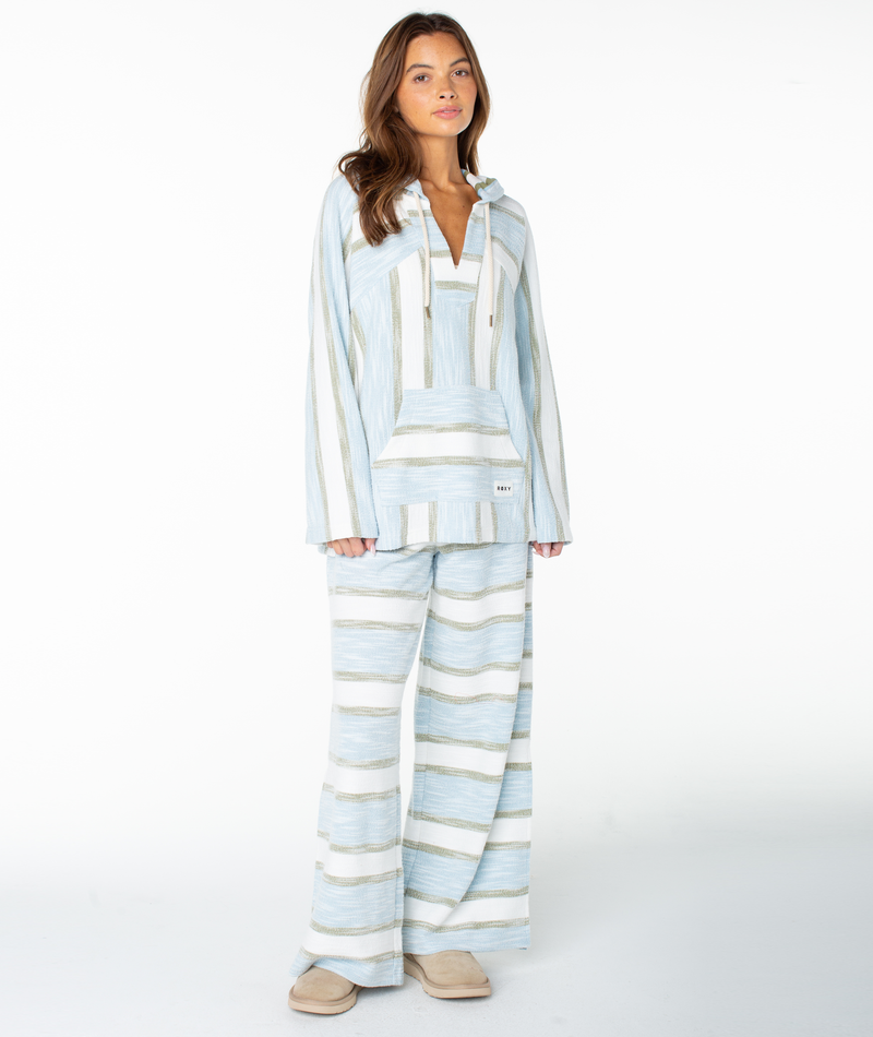 A woman wears the Roxy Wild And Free Top, a loose-fitting light blue and white striped hoodie crafted from a soft cotton blend terry, paired with matching wide-leg pants. Standing on a white background in beige slippers, she effortlessly channels relaxed coastal vibes.